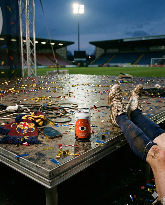 Brulo King for a Day can on a confetti-covered stage after a football match, with a fan scarf and a player's muddy boots