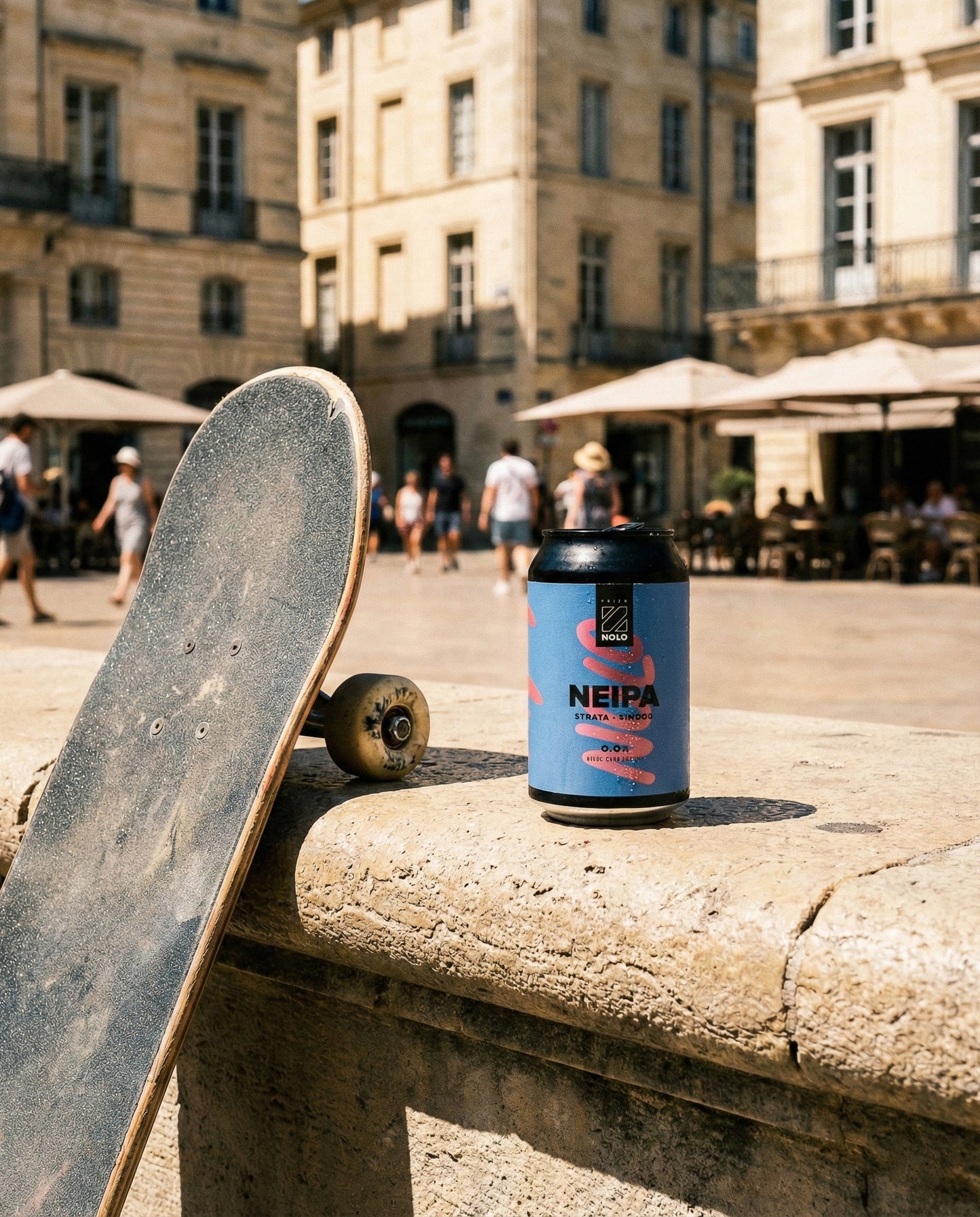 Prizm NOLO NEIPA can on a stone bench in a sun-baked southern French city plaza with a skateboard leaning beside it